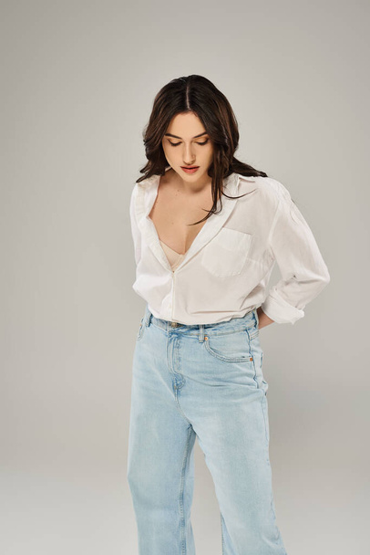 Krásná plus velikost žena pózuje sebevědomě ve stylové bílé košili a modré džíny na šedém pozadí. - Fotografie, Obrázek