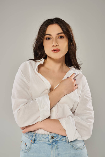 Stylish plus-size woman posing in trendy white shirt and jeans against a gray backdrop. - Photo, Image