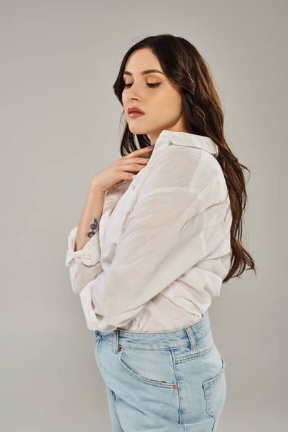 A stunning plus-size woman poses elegantly in a white shirt and jeans against a gray backdrop. - Photo, Image