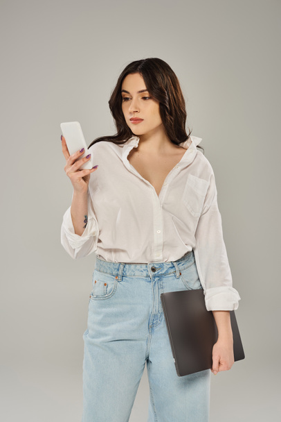 Een stijlvolle plus-size vrouw poseert in een wit shirt, vol vertrouwen met een mobiele telefoon tegen een grijze achtergrond. - Foto, afbeelding