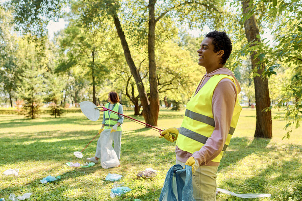 Una pareja cariñosa y diversa en chalecos de seguridad y guantes limpian un parque juntos, de pie en la exuberante hierba verde. - Foto, Imagen