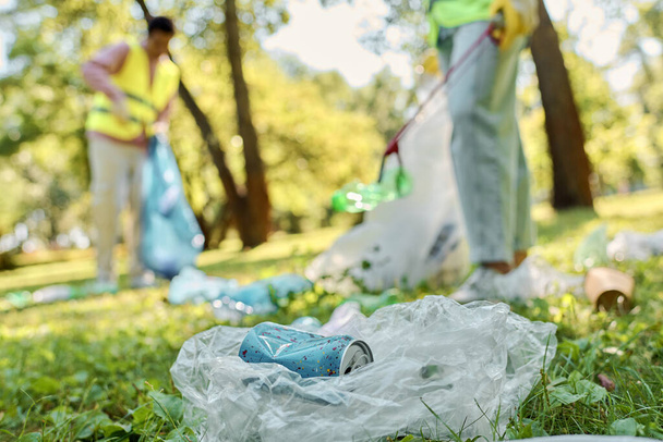 Ένα κοινωνικά ενεργό, ποικιλόμορφο ζευγάρι με γιλέκα και γάντια ασφαλείας που καθαρίζει τα σκουπίδια σε ένα πάρκο, προωθώντας την προστασία του περιβάλλοντος και τη συμμετοχή της κοινότητας. - Φωτογραφία, εικόνα