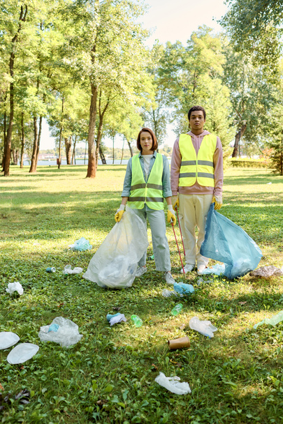 Una coppia socialmente attiva diversificata in giubbotti di sicurezza e guanti in piedi insieme nella lussureggiante erba verde, pulire il parco. - Foto, immagini