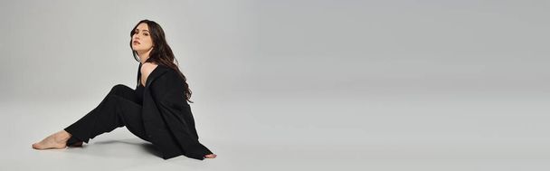 Een mooie vrouw van plus-size zit sierlijk op de grond in een stijlvolle zwarte outfit tegen een grijze achtergrond. - Foto, afbeelding