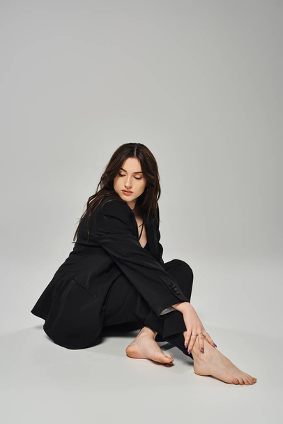 Eine schöne Plus-Size-Frau in einem eleganten schwarzen Mantel sitzt anmutig auf dem Boden vor grauem Hintergrund. - Foto, Bild