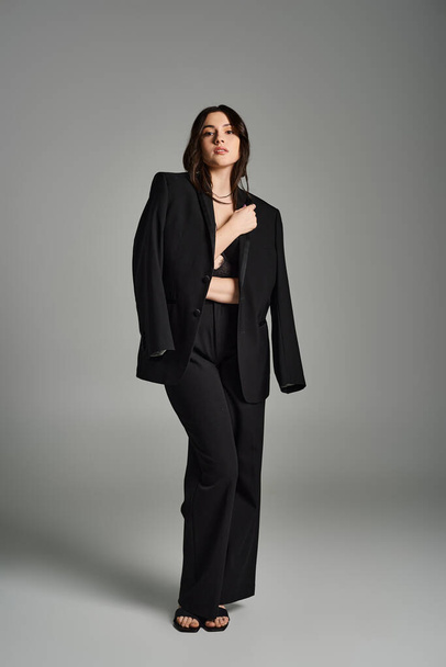 Ошеломляющая плюс размер женщина демонстрирует свою элегантность в шикарном черном костюме на гладком сером фоне. - Фото, изображение