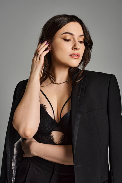 A beautiful plus-size woman exudes confidence in a black suit and bra against a gray backdrop. - Photo, Image