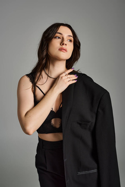 Stylish plus size woman confidently poses in a black suit and bra against a gray backdrop. - Photo, Image