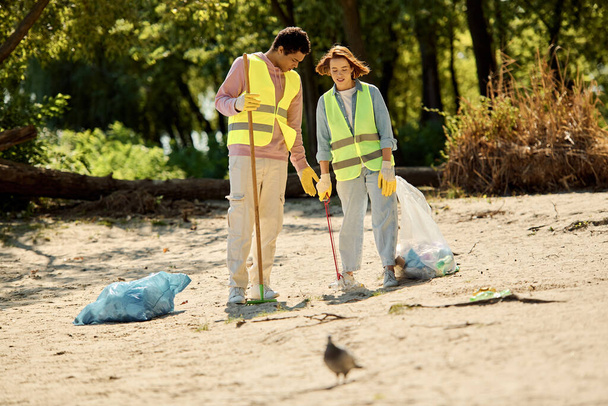 Una pareja diversa, con chalecos y guantes de seguridad, limpian un parque mientras están parados en la arena juntos. - Foto, imagen