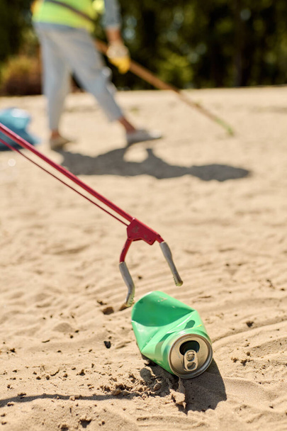 Zelená plechovka s červenou rukojetí sedí na písečné pláži, symbolizující péči o životní prostředí a úklid pláže. - Fotografie, Obrázek