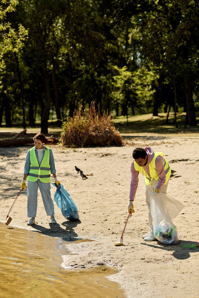 Un couple socialement actif et diversifié en gilets de sécurité et gants, debout dans le sable, nettoyant le parc ensemble. - Photo, image