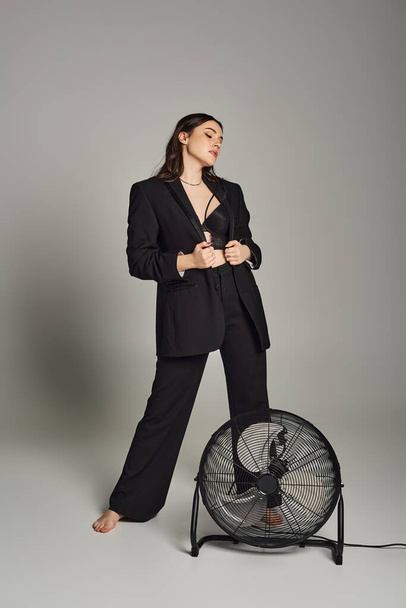 Una mujer elegante de talla grande emana confianza en un traje a medida, de pie con gracia junto a un ventilador giratorio en un fondo gris. - Foto, imagen