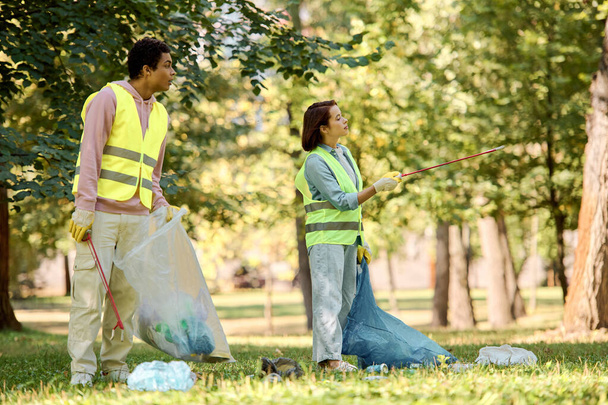 A loving, diverse couple in safety vests and gloves stand side by side on lush green grass, engaged in park cleanup. - Photo, Image