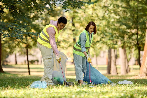 A socially active, diverse couple in safety vests and gloves stands united in the lush grass, passionately cleaning the park. - Photo, Image