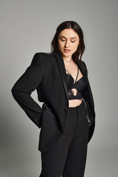 A stunning plus size woman in a sleek black suit strikes a confident pose against a gray backdrop. - Foto, imagen