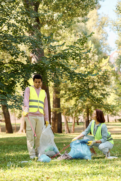 Una coppia diversificata in giubbotti di sicurezza e guanti in piedi nell'erba, pulire un parco insieme. - Foto, immagini