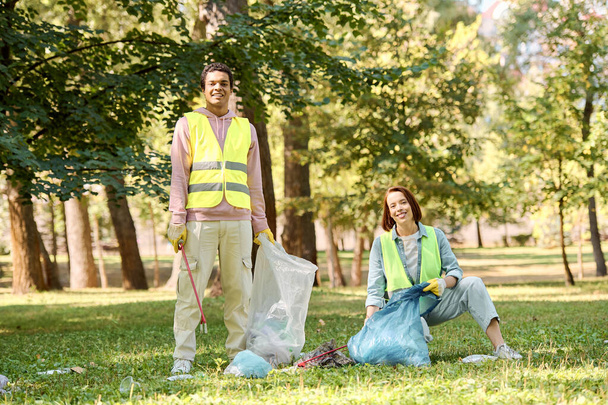 Разнообразная пара в защитных жилетах и перчатках очищает парк, стоящий в траве, объединенный в своей преданности окружающей среде. - Фото, изображение