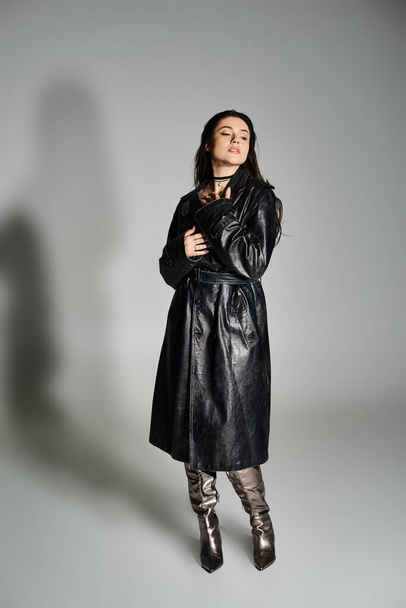 Eine atemberaubende Plus-Size-Frau posiert in schickem schwarzen Mantel und Stiefeln vor grauem Hintergrund. - Foto, Bild
