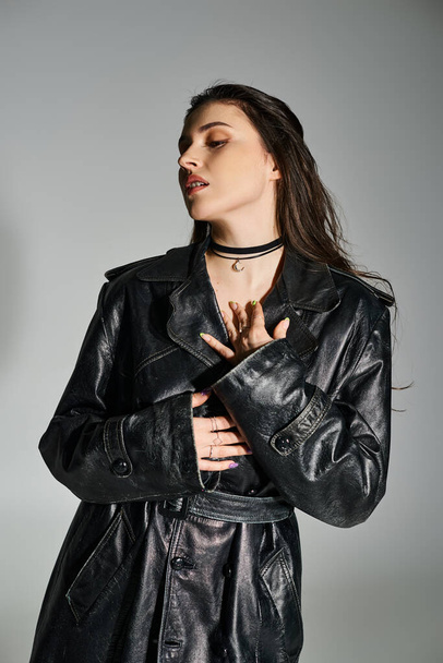 Een mooie vrouw met een extra maat slaat een pose in een strak zwart leren jasje en trendy choker tegen een grijze achtergrond. - Foto, afbeelding