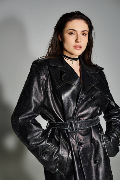 Eine atemberaubende Plus-Size-Frau strahlt in einem schwarzen Leder-Trenchcoat vor neutralem grauen Hintergrund Zuversicht aus. - Foto, Bild