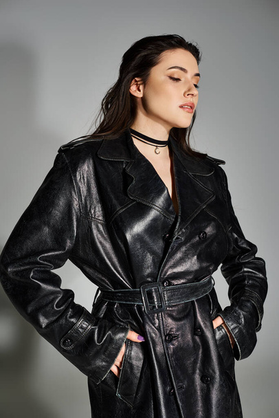 Uma mulher deslumbrante plus size marcando uma pose em um casaco de trincheira de couro preto contra um pano de fundo cinza. - Foto, Imagem