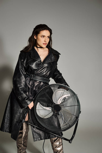 Una hermosa mujer de talla grande posando en un abrigo de cuero de moda, sosteniendo un ventilador, exudando confianza y estilo en un fondo gris. - Foto, Imagen