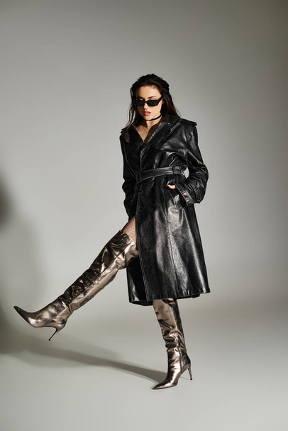 Een mooie plus size vrouw poseert zelfverzekerd in een stijlvolle zwarte jas en bijpassende laarzen tegen een grijze achtergrond. - Foto, afbeelding