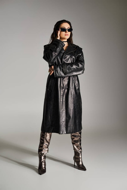 Eine Plus-Size-Frau strahlt in stylischem schwarzen Ledermantel und Stiefeln vor grauem Hintergrund Zuversicht aus. - Foto, Bild