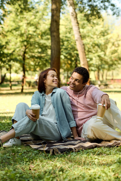 Zróżnicowana para siedzi na kocu w parku, ciesząc się swoim towarzystwem w żywym stroju. - Zdjęcie, obraz