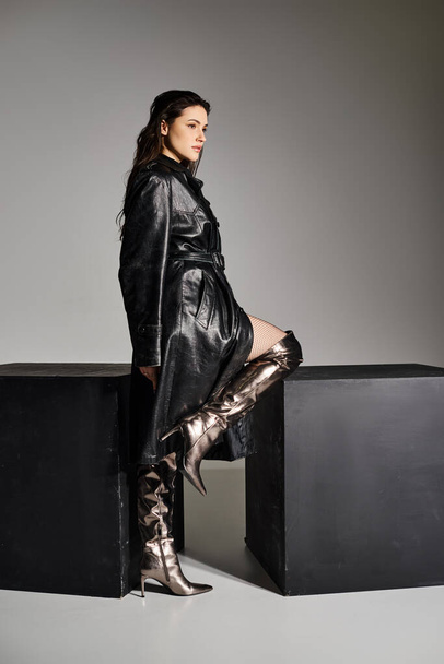 Eine schöne Plus-Size-Frau in stylischer Kleidung posiert auf einem schwarzen Kasten vor grauem Hintergrund. - Foto, Bild