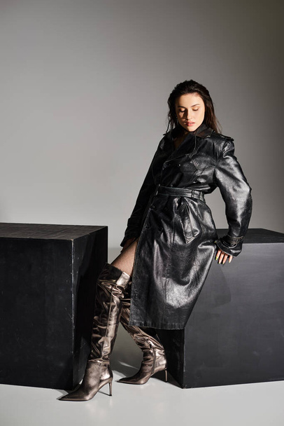 Μια γυναίκα συν μέγεθος με μαύρο παλτό και μπότες ακουμπά σε ένα μαύρο κουτί σε γκρι φόντο, αποπνέοντας κομψότητα και αυτοπεποίθηση. - Φωτογραφία, εικόνα