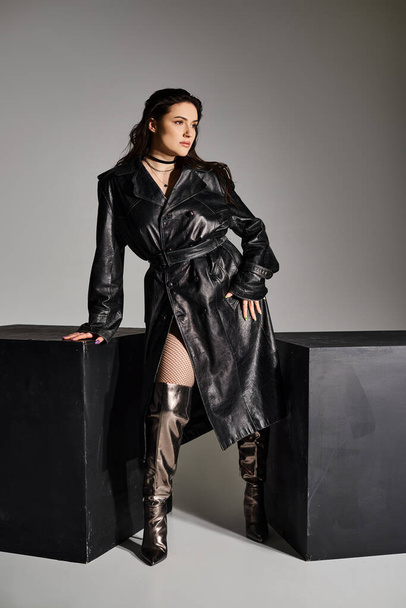 Приголомшлива жінка плюс розмір, що демонструє свій стиль в чорному траншеї пальто і чоботи на сірому тлі. - Фото, зображення