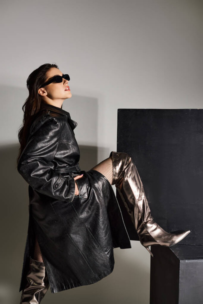 Uma bela mulher plus-size posa confiantemente em um casaco preto e botas de prata contra um pano de fundo cinza. - Foto, Imagem