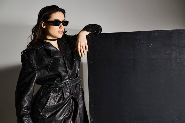 Une femme élégante plus taille frappe une pose dans un manteau en cuir noir et des lunettes de soleil sur fond gris. - Photo, image