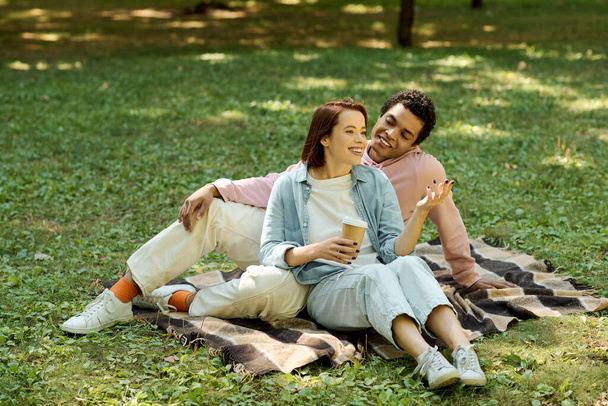 Um casal diversificado vestido com trajes vibrantes se senta em um cobertor na grama, desfrutando de um momento de paz juntos no parque. - Foto, Imagem