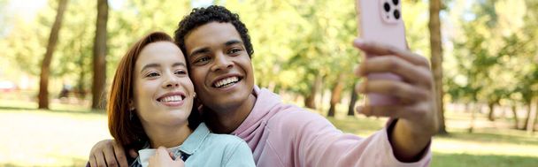 Un couple aimant et diversifié, vêtu vibramment, capturant un moment heureux ensemble alors qu'ils prennent un selfie dans un parc. - Photo, image