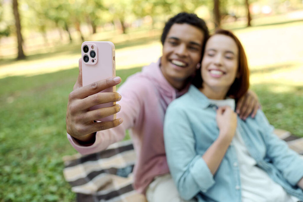 Un couple aimant et diversifié en tenue vibrante capture un moment de bonheur ensemble en prenant un selfie dans un magnifique cadre de parc. - Photo, image