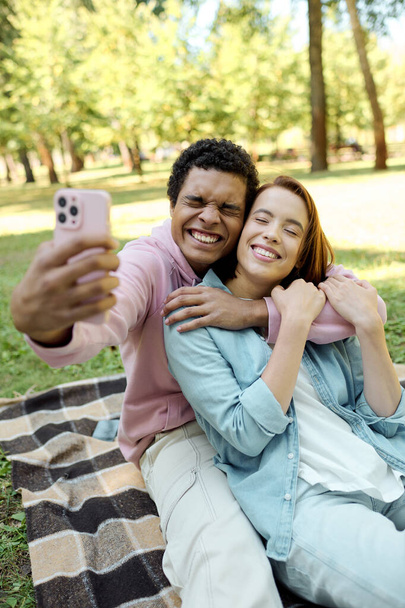 Ein Mann in lebendiger Kleidung macht ein Selfie mit einer Frau auf einer Decke im Park und genießt einen liebevollen Moment miteinander. - Foto, Bild