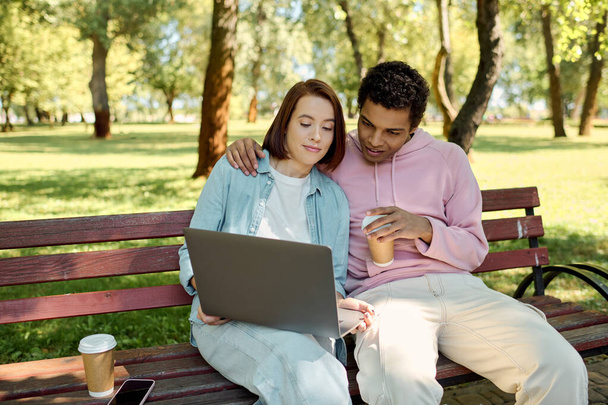 A man and woman in vibrant attire sit on a park bench, engrossed in a laptop screen, enjoying quality time together outdoors. - Photo, Image