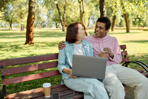 Ένα ποικίλο ζευγάρι με ζωντανή ενδυμασία κάθεται σε ένα παγκάκι του πάρκου, χρησιμοποιώντας ένα φορητό υπολογιστή μαζί, βυθισμένο στον ψηφιακό κόσμο τους, ενώ περιβάλλεται από τη φύση. - Φωτογραφία, εικόνα