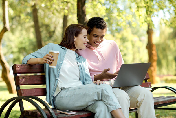 Tyylikäs pari istuu puistossa penkki, engrossed kannettavan tietokoneen näytön, nauttia laatuaikaa vilkkaassa ympäristössä. - Valokuva, kuva