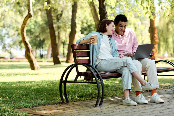 Ένα ζευγάρι με έντονα ρούχα κάθεται σε ένα παγκάκι, απορροφημένο στο laptop τους. - Φωτογραφία, εικόνα