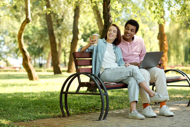 A diverse couple sitting on a park bench, working together on a laptop, surrounded by nature. - Фото, изображение