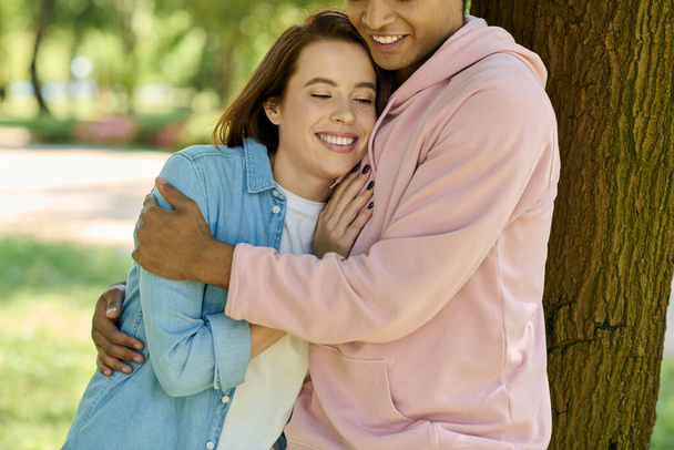 Um homem e uma mulher de traje vibrante abraçam ternamente na frente de uma árvore majestosa em um parque, mostrando sua profunda conexão. - Foto, Imagem