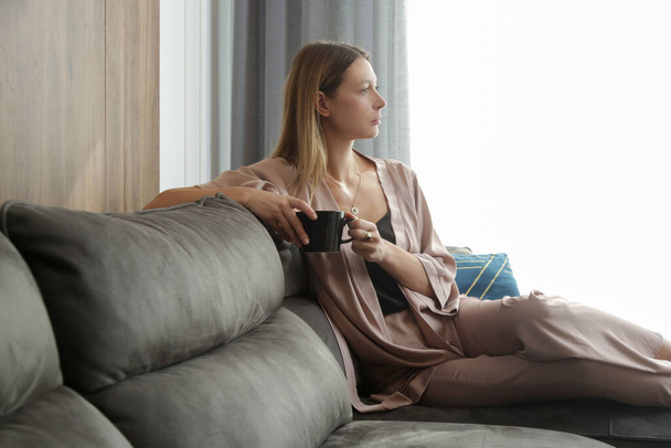 Mujer en ropa de satén tomando una taza de té, descansando y sentada en la sala de estar - Foto, Imagen