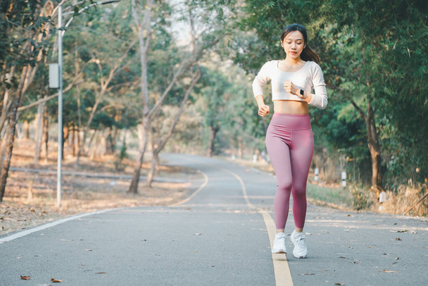 Fitness koncept, Soustředěná mladá běžkyně s fitness trackerem a sluchátky běžícími po stezce parku obklopená stromy - Fotografie, Obrázek