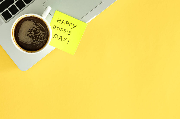 Tahmea huomautus lauseella Happy Boss Day kiinnitetty kannettavan tietokoneen näppäimistöön ja kuppi kahvia keltaisella toimistopöydällä, ylhäältä. - Valokuva, kuva