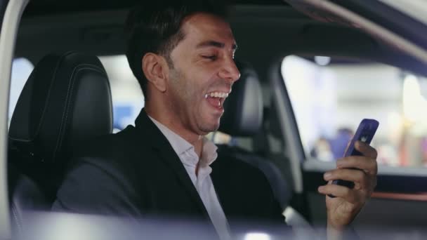 成功したビジネスマンは彼の車に座って,面白いコンテンツをスクロールし,彼のスマートフォンで入力ビデオを楽しんでいます. 男性の笑顔の車の所有者はビデオを見て笑っています. テクノロジーとビジネス - 映像、動画