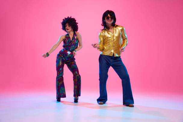 Чоловік і жінка, одягнені в яскраве вбрання 1970-х років, танцюють у русі проти градієнтного рожевого студійного фону. Ретро мода. Концепція американської культури, 1970-х, 1980-х років мода, музика, порівняння епох. - Фото, зображення