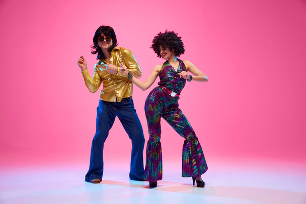 Grooviges Tanzpaar in hellem Gewand, das Disco-Bewegungen in Bewegung vor gradienten rosafarbenen Studiohintergrund vollführt. Konzept der amerikanischen Kultur, Mode der 1970er, 1980er Jahre, Musik, Epochenvergleiche. - Foto, Bild